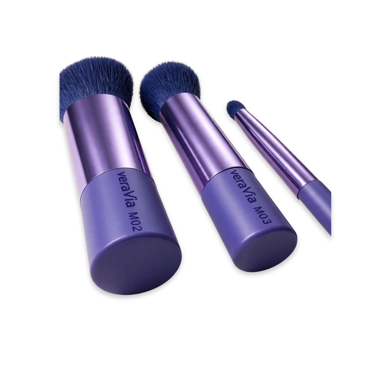 Three veraVia travel brushes upright at an angle showing labelled handles: M02 with a wide flat-top navy bristle head, M03 with a rounded dome head, and a slim M04 precision brush — all on purple metallic handles with matte purple end caps