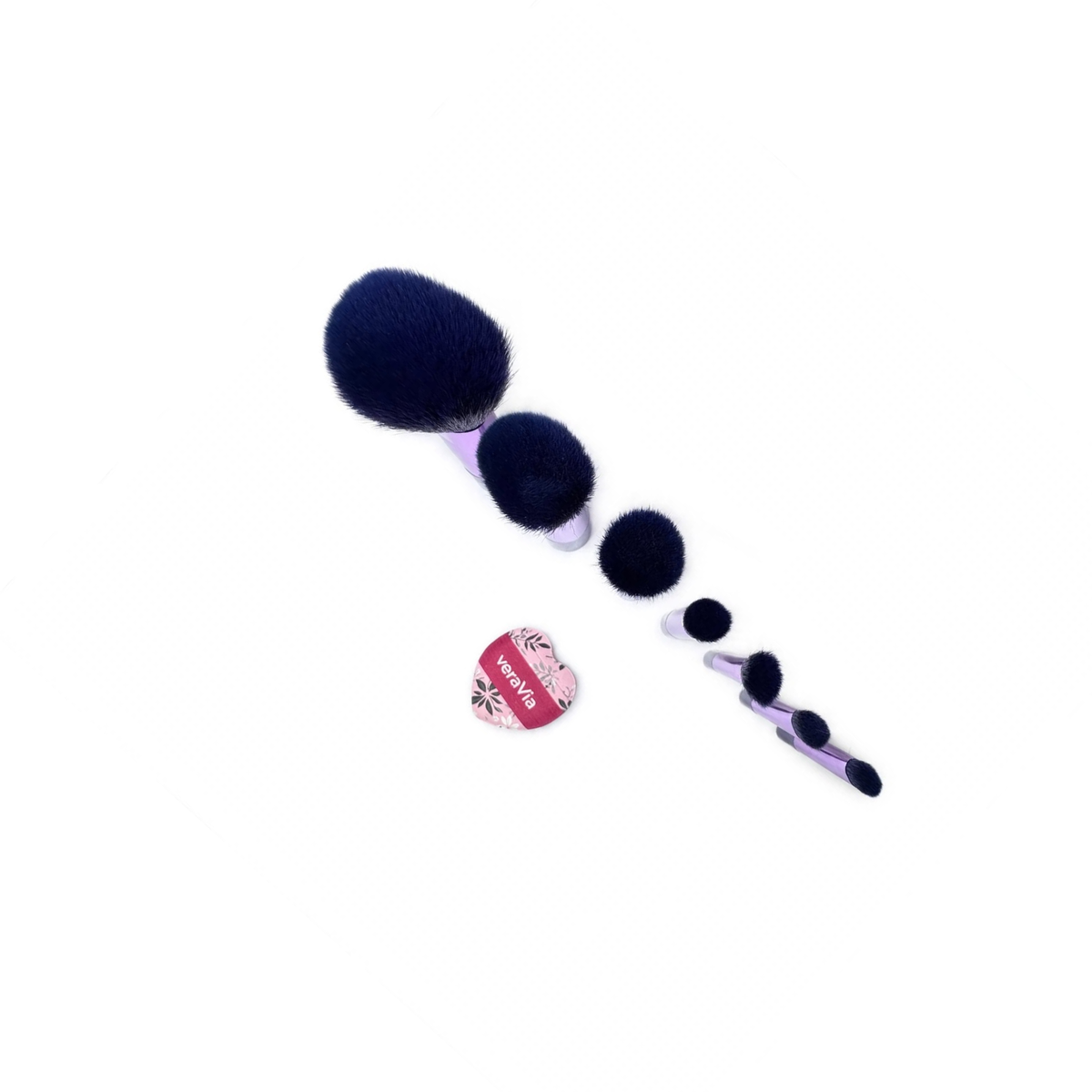 Diagonal top-down view of seven travel makeup brush heads in navy bristles decreasing from a large dome to a tiny flat tip, with a heart-shaped veraVia mini puff centered separately below the brush cluster, on white background. Designed for travel, it’s portable and compact.
