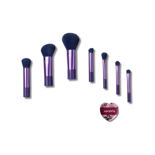 Seven veraVia travel brushes with navy bristle heads and purple metallic handles arranged in a rising diagonal arc by size, with a heart-shaped veraVia-branded mini air cushion puff at bottom right, on white background, especially for travel, portable and mini.
